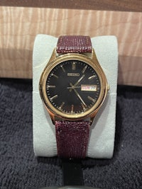 a watch with a gold band and burgundy strap sitting on top of a box